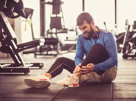 Man holding his injured ankle while sitting on the floor of a gym